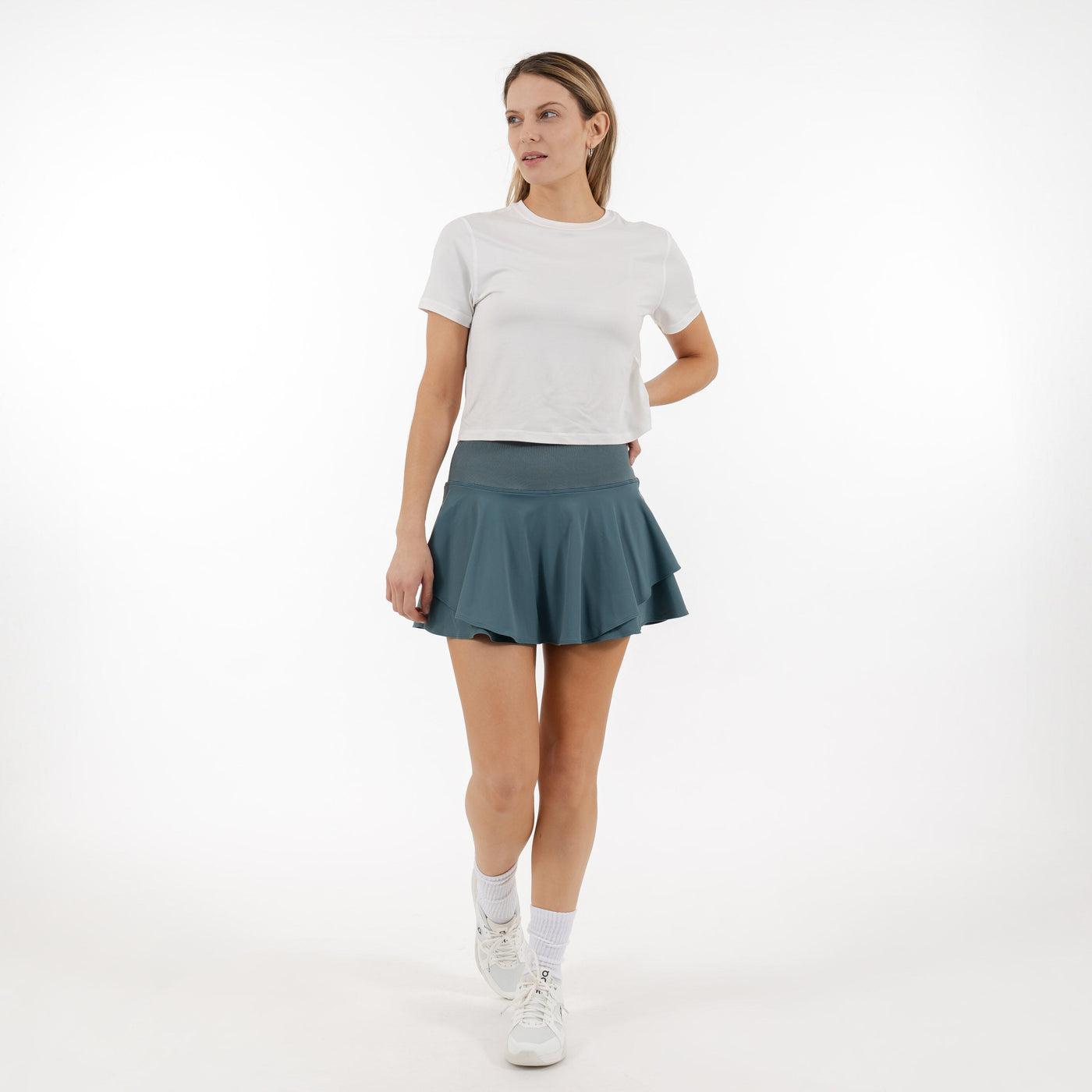 A woman poses against a white background, wearing a white tee and the RHOBACK Sydney Tennis Skort | Solid - Jungle Green with a high-rise ribbed waistband. She looks to the side, her skort crafted from moisture-wicking fabric.