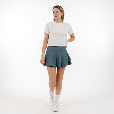 A woman poses against a white background, wearing a white tee and the RHOBACK Sydney Tennis Skort | Solid - Jungle Green with a high-rise ribbed waistband. She looks to the side, her skort crafted from moisture-wicking fabric.