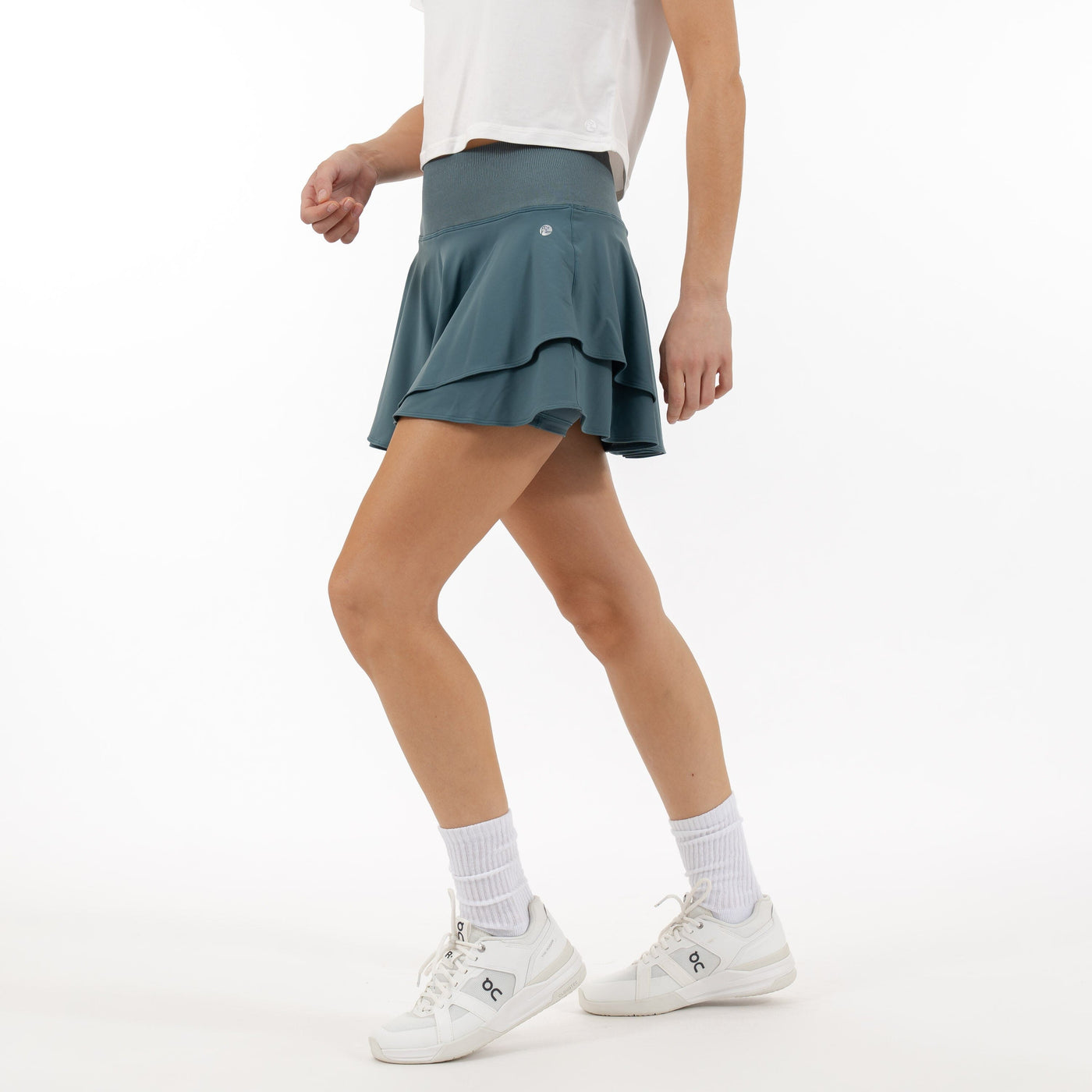 A person in a white shirt and RHOBACK Sydney Tennis Skort | Solid - Jungle Green with layered ruffles and a high-rise ribbed waistband walks forward, pairing it with white socks and sneakers against a plain white background.