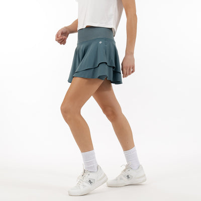 A person in a white shirt and RHOBACK Sydney Tennis Skort | Solid - Jungle Green with layered ruffles and a high-rise ribbed waistband walks forward, pairing it with white socks and sneakers against a plain white background.