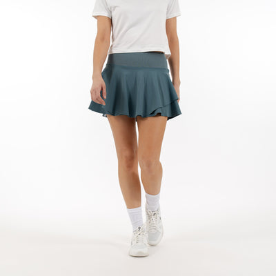 A person wearing a fitted white t-shirt and the RHOBACK Sydney Tennis Skort | Solid - Jungle Green, with a high-rise ribbed waistband, white crew socks, and white sneakers stands against a plain white background. Their face is not visible.