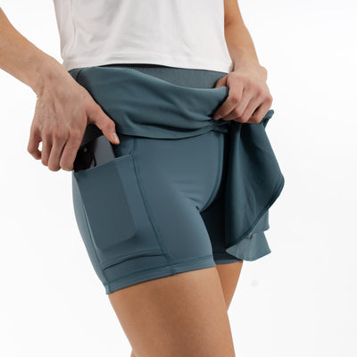 A person wearing the RHOBACK Sydney Tennis Skort in Jungle Green with a high-rise ribbed waistband and phone pocket holds up a matching jacket, shown from the waist down against a white background.