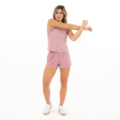 A woman in the RHOBACK Drift Performance Crop Tank, Heather - Dusk Pink/Lotus Pink, stretches her right arm across her chest against a plain white background. She pairs it with shorts and white sneakers.