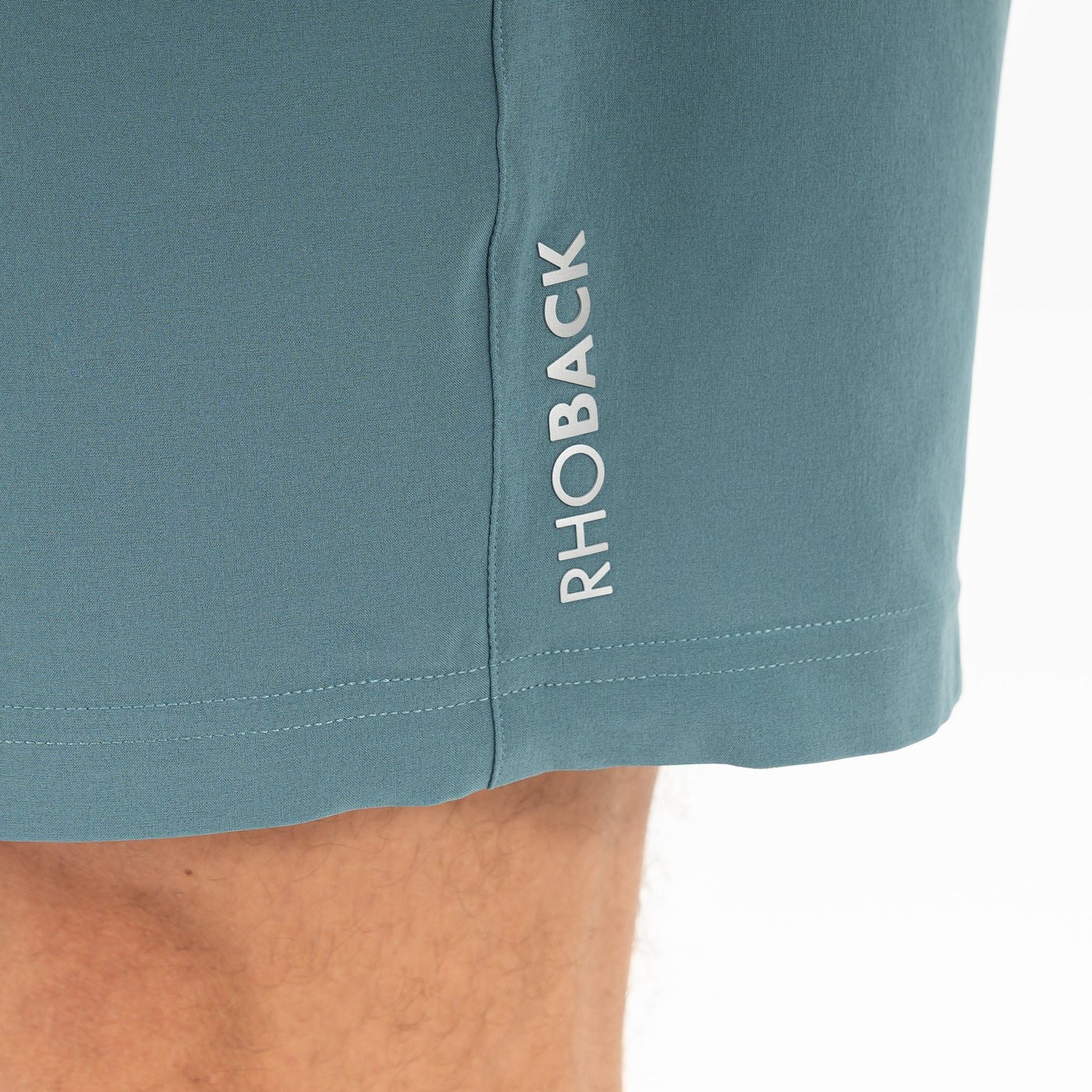 Close-up of RHOBACK All-Active Short | Solid - Jungle Green, made from lightweight, quick-drying fabric with "RHOBACK" printed vertically in white near the hem. A part of a person's leg appears at the bottom left.