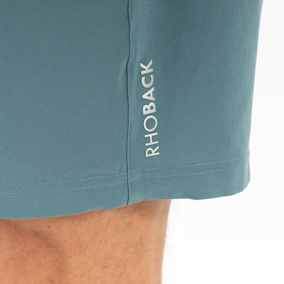 Close-up of RHOBACK All-Active Short | Solid - Jungle Green, made from lightweight, quick-drying fabric with "RHOBACK" printed vertically in white near the hem. A part of a person's leg appears at the bottom left.