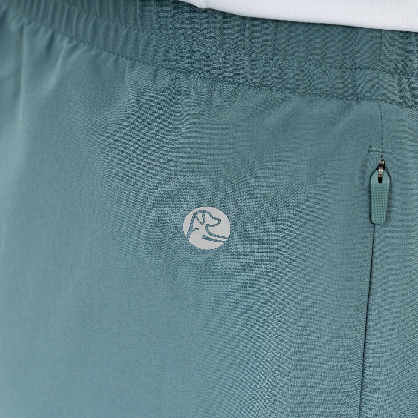 Close-up of RHOBACK All-Active Short in Jungle Green, crafted from quick-drying, lightweight fabric with an elastic waistband, zippered side pocket, and a small circular logo featuring a dog’s head outline.
