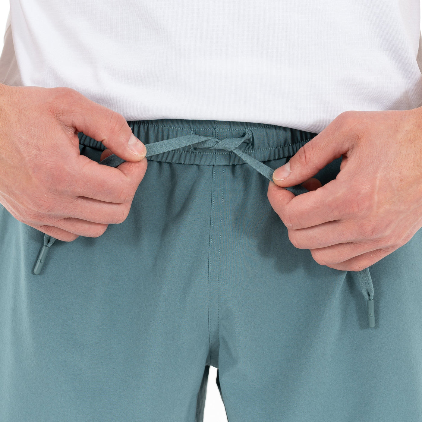 Close-up of hands tying the drawstring on RHOBACK All-Active Short | Solid - Jungle Green, made from quick-drying, lightweight fabric. Person wears a white shirt; only their hands, torso, and the shorts are visible.