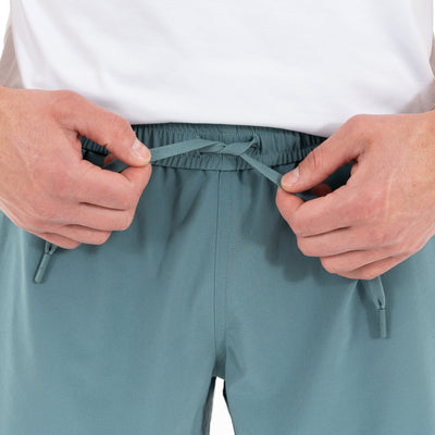Close-up of hands tying the drawstring on RHOBACK All-Active Short | Solid - Jungle Green, made from quick-drying, lightweight fabric. Person wears a white shirt; only their hands, torso, and the shorts are visible.