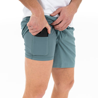 A person, shown from the waist down against a white background, places a smartphone into the side pocket of RHOBACK’s All-Active Short | Solid in Jungle Green, paired with a white shirt.