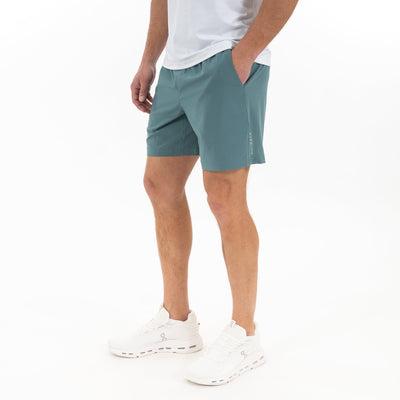 A person wears a white t-shirt, RHOBACK All-Active Short | Solid in Jungle Green, and white sneakers, standing with hands in pockets against a plain white background.