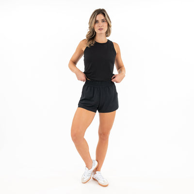 A woman poses against a white background in RHOBACK's Era High-Rise Short | Solid - Midnight Black, a sleeveless black top, and white sneakers, one hand on her hip and one leg slightly bent.