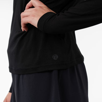 A person wears the RHOBACK Drift Performance Long Sleeve Tee | Solid - Midnight Black, made from wrinkle-resistant SkyKnit™ fabric, with black pants. Only their midsection and arm are visible against a plain white background.