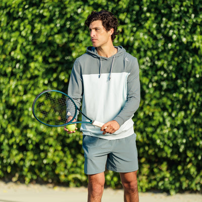 A young man wears the RHOBACK Hesi Performance Hoodie | Colorblock - Jungle Green/Aquafrost and shorts, holding a tennis racket and ball on an outdoor court with a green leafy background.