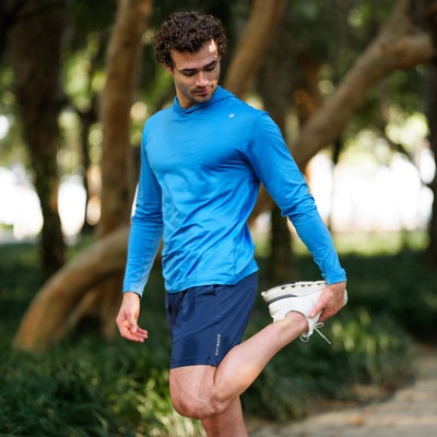 A man in the RHOBACK Blitz Tech Hoodie in Heather - Birdie Blue/Sapphire Blue stretches his leg outdoors on a path surrounded by trees, wearing navy shorts with moisture-wicking SilverKnit fabric.