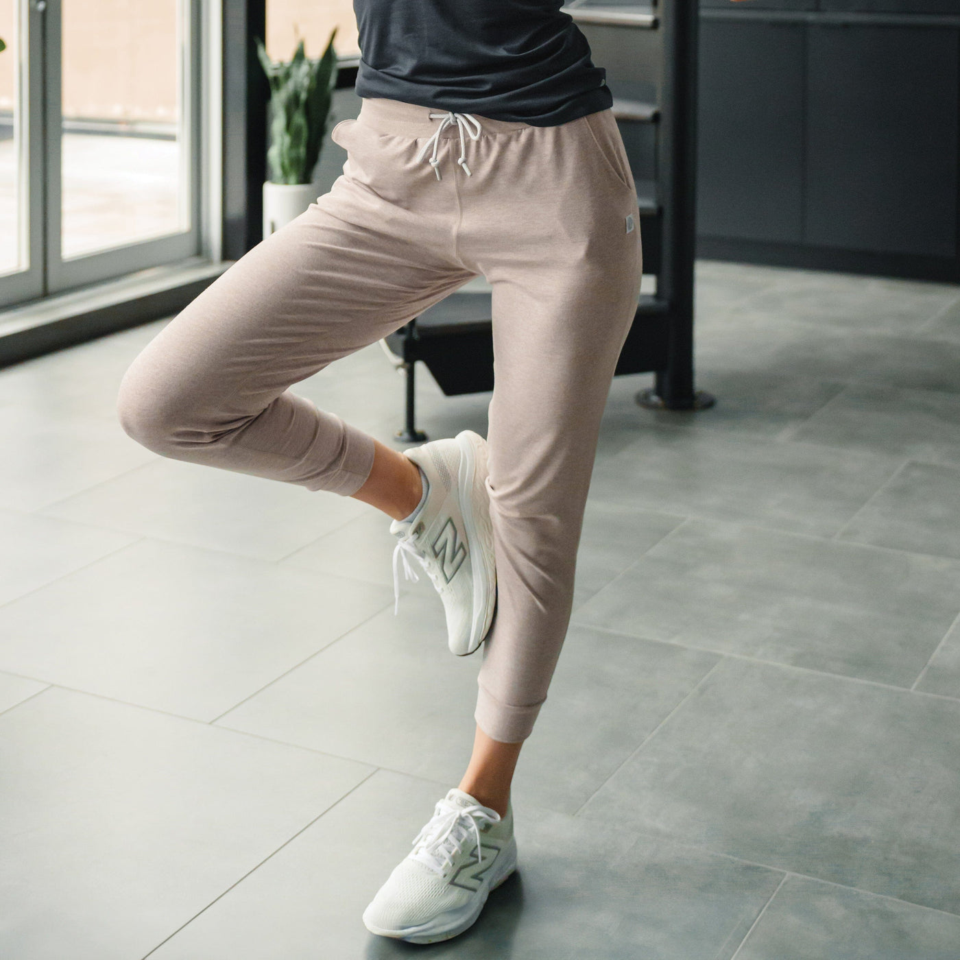 A person wearing RHOBACK's Tempo Performance Jogger in Heather - Warm Stone/White, made with four-way stretch, stands indoors on one leg in a yoga pose on a tiled floor with natural light from large windows.