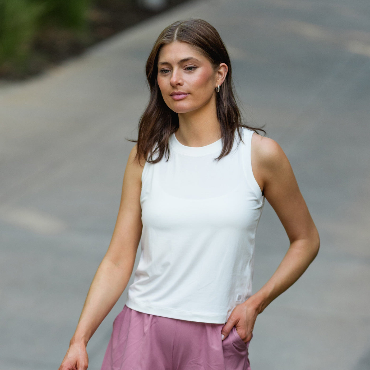 A woman with straight brown hair walks outside in the RHOBACK Drift Performance Crop Tank | Solid - White, pairing it with pink shorts. With one hand in her pocket and a calm expression, she enjoys the greenery along a paved path.
